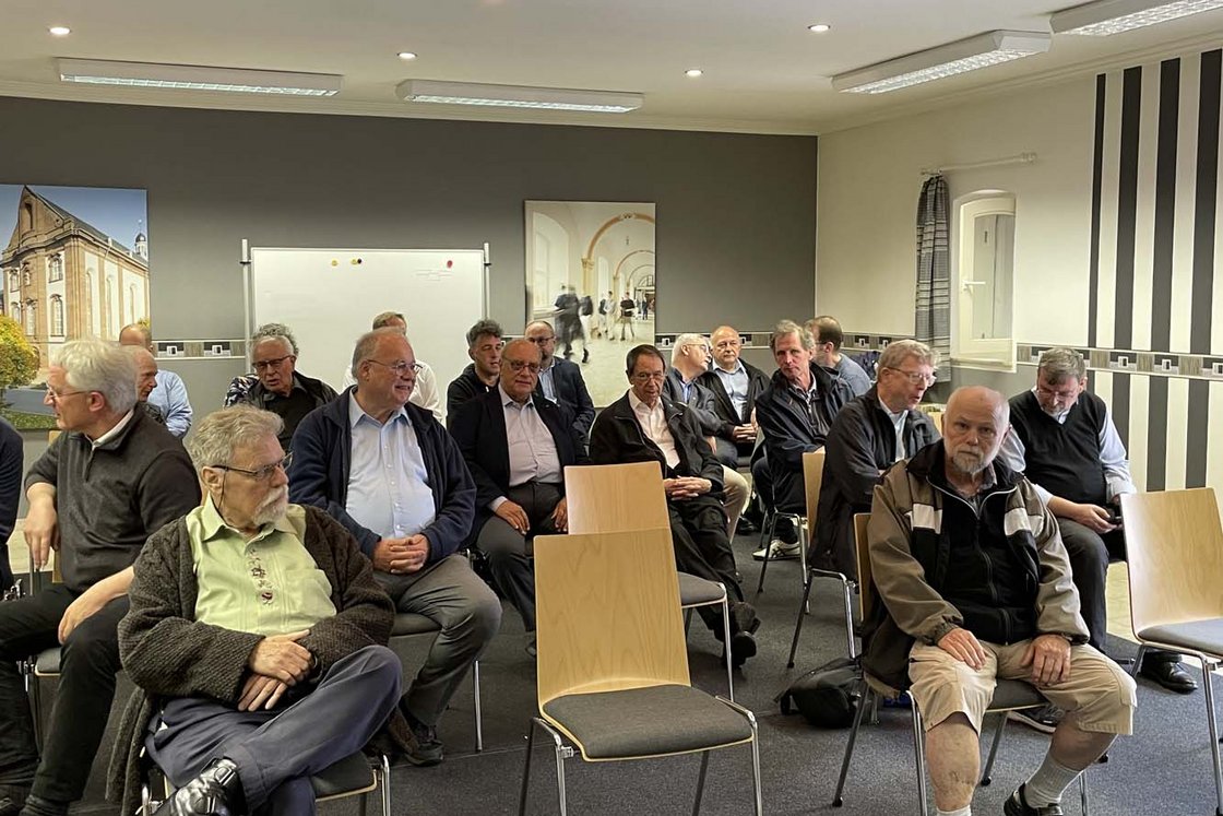 Eine Gruppe älterer Männer sitzt in Reihen auf Stühlen und schaut aufmerksam nach vorne in einem hell erleuchteten Raum.