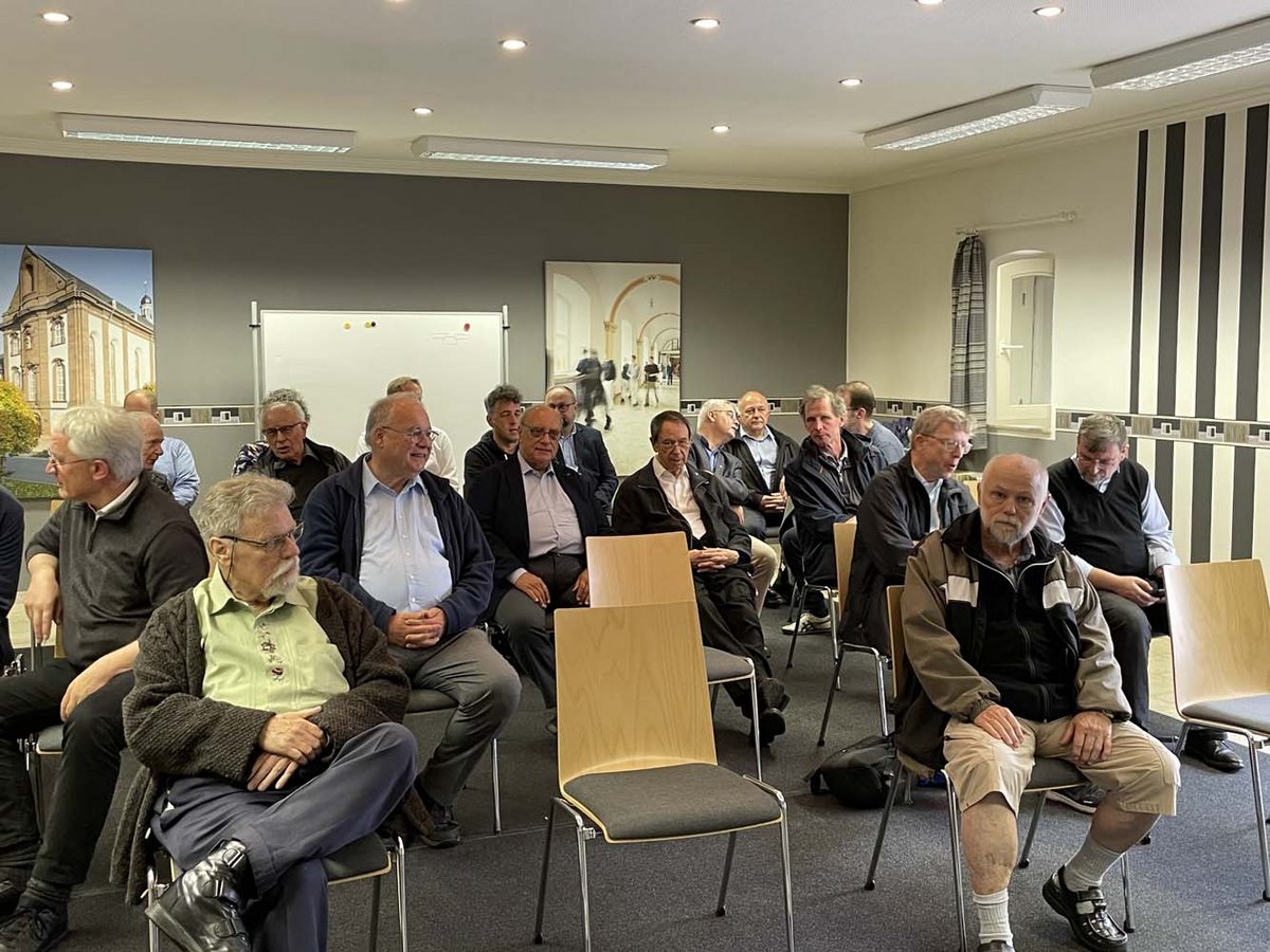 Eine Gruppe älterer Männer sitzt in Reihen auf Stühlen und schaut aufmerksam nach vorne in einem hell erleuchteten Raum.