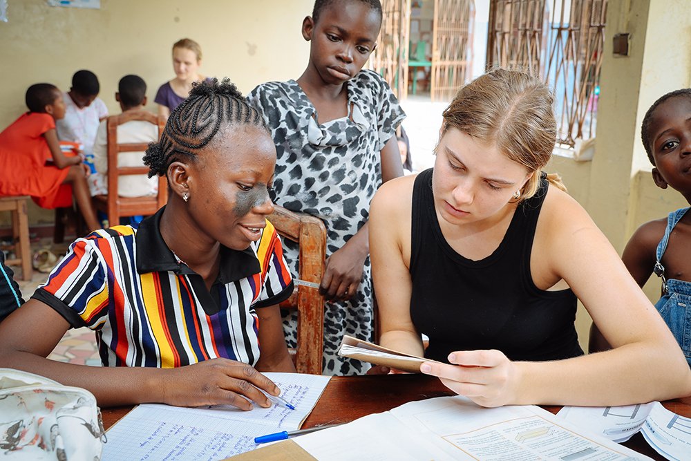 Zwei junge Frauen lernen gemeinsam an einem Tisch, eine schreibt, die andere liest aus einem Buch.