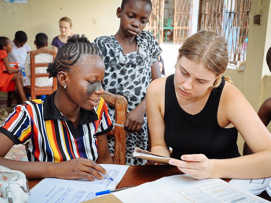 Zwei junge Frauen lernen gemeinsam an einem Tisch, eine schreibt, die andere liest aus einem Buch.