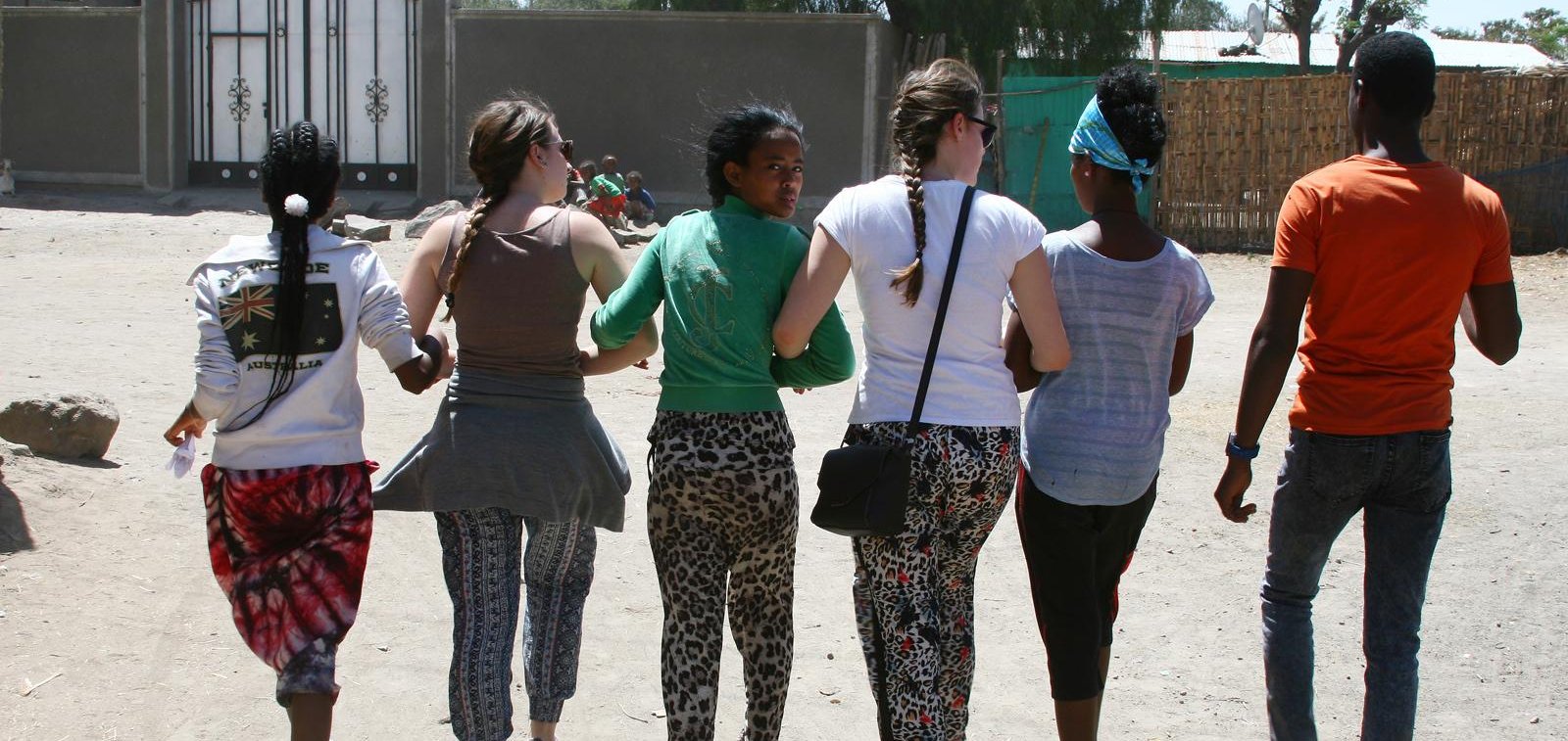 Sechs junge, vielfältige Menschen gehen Arm in Arm auf einem staubigen Weg, von hinten gesehen, einer blickt zurück.