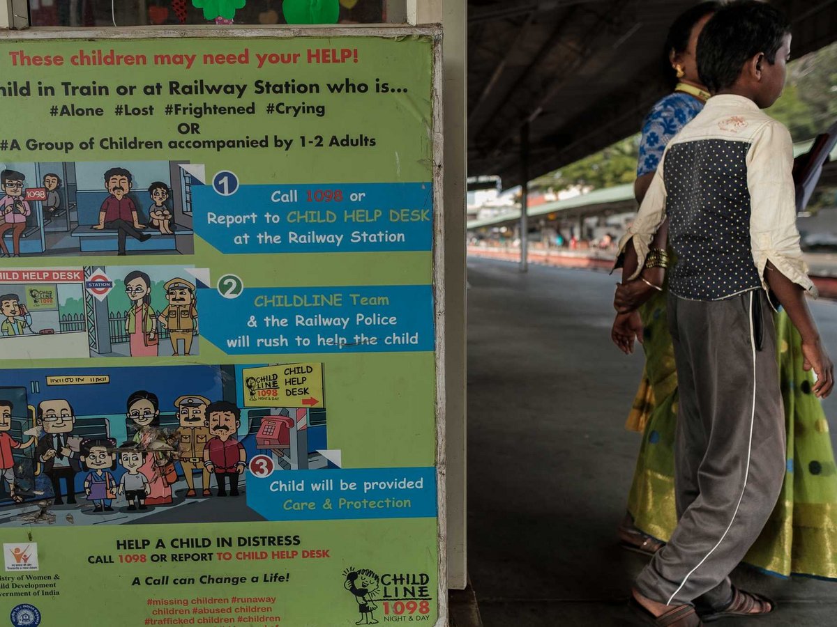 Schild für Kinderhilfe an indischem Bahnhof mit Anweisungen. Unscharf gehen ein Junge und eine Frau daneben auf dem Bahnsteig.