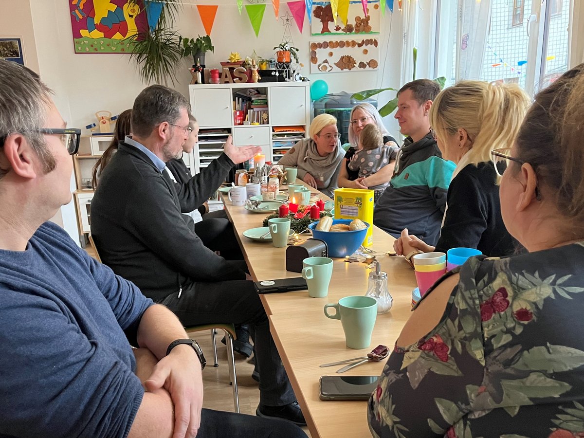 Eine Gruppe Erwachsener und ein Baby sitzen an einem dekorierten Tisch mit Snacks und Getränken, unterhalten sich.