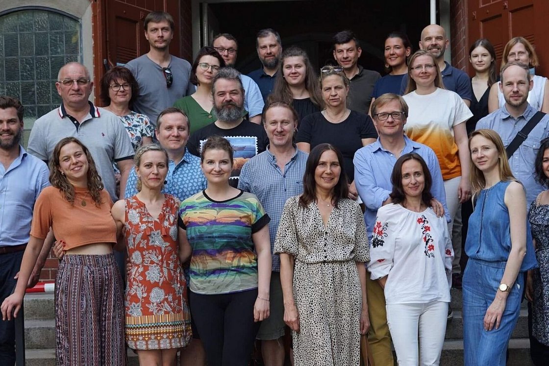 Eine vielfältige Gruppe erwachsener Männer und Frauen posiert lächelnd für ein Gruppenfoto auf den Stufen eines Ziegelgebäudes.