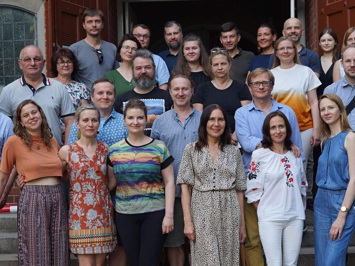 Eine vielfältige Gruppe erwachsener Männer und Frauen posiert lächelnd für ein Gruppenfoto auf den Stufen eines Ziegelgebäudes.