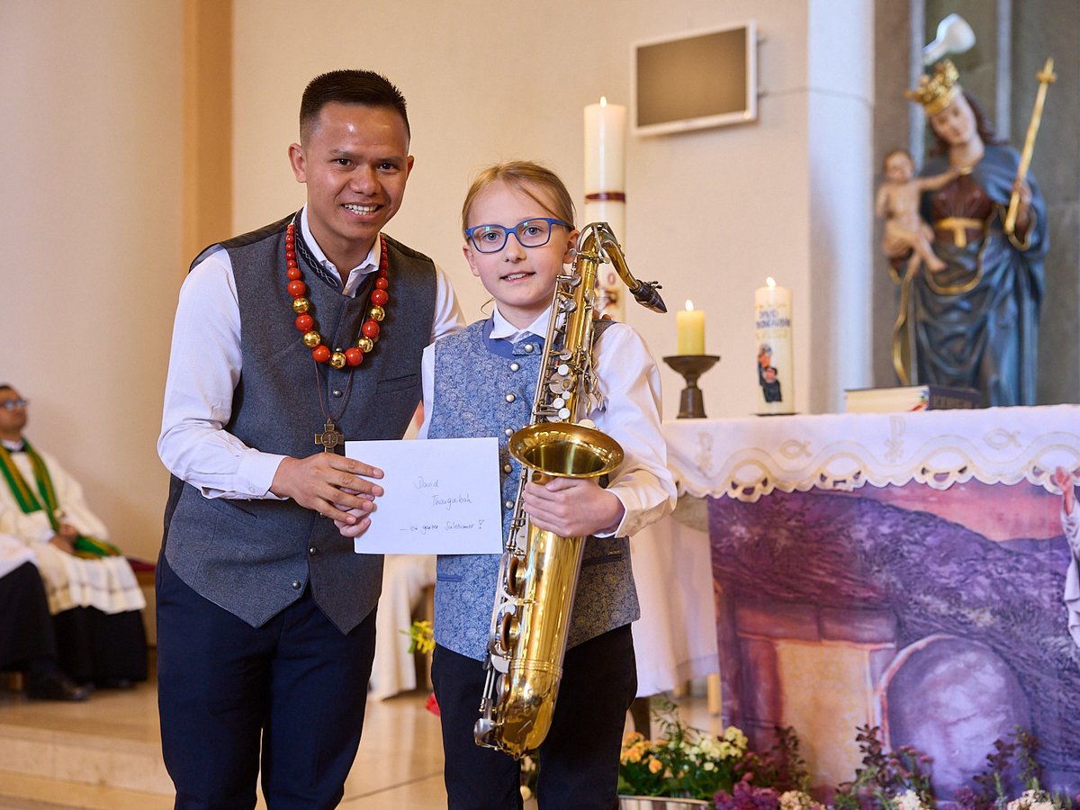 Ein Mann und ein Mädchen lächeln in einer Kirche. Das Mädchen hält ein goldenes Saxophon, der Mann ein Dokument.