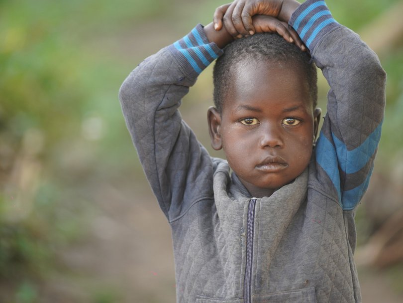 Ein kleines Kind mit dunkler Haut und ernsten Augen, Hände auf dem Kopf verschränkt, trägt eine graue Jacke vor grünem Hintergrund.