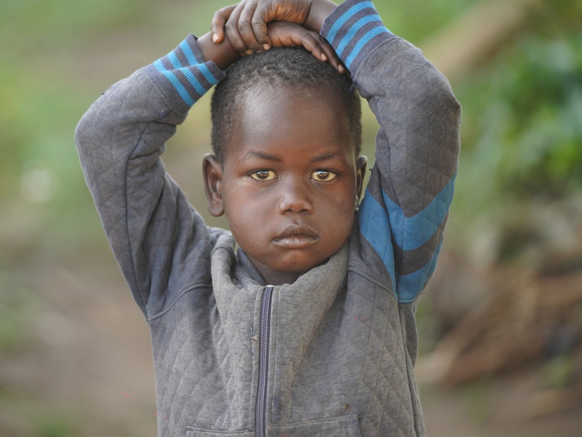 Ein kleines Kind mit dunkler Haut und ernsten Augen, Hände auf dem Kopf verschränkt, trägt eine graue Jacke vor grünem Hintergrund.