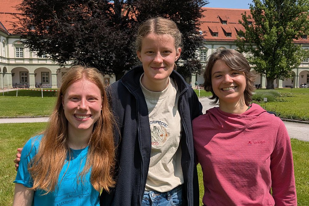 Drei lächelnde junge Frauen stehen an einem sonnigen Tag im Freien vor einem historischen Gebäude mit Bögen und Bäumen.