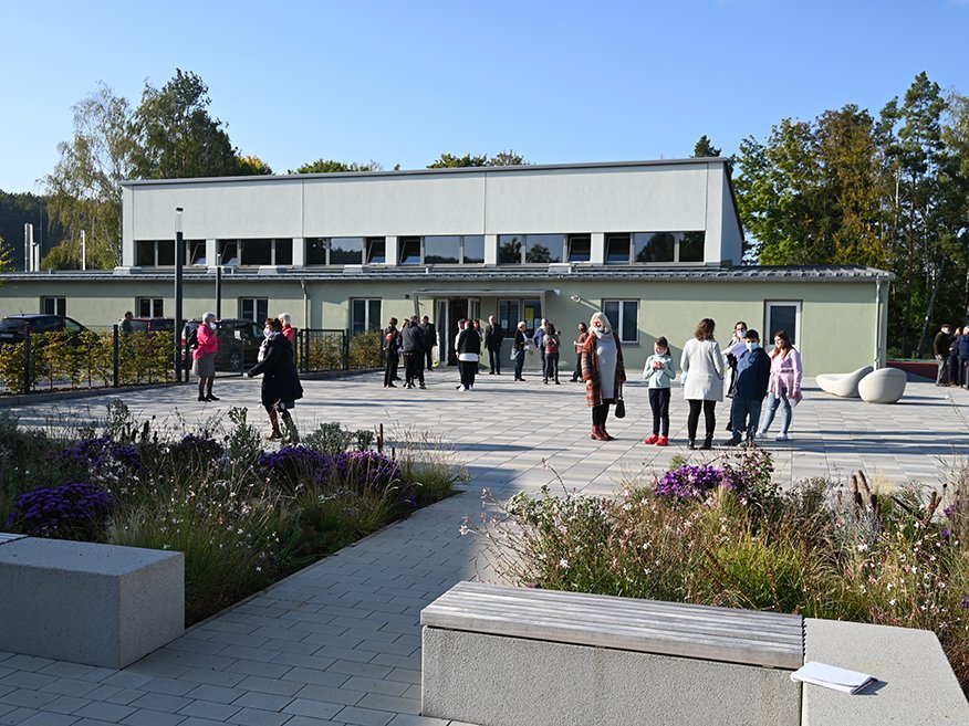 Gepflasterter Vorplatz mit Menschen vor einem modernen Gebäude, umgeben von Grünanlagen und Bänken an einem sonnigen Tag.