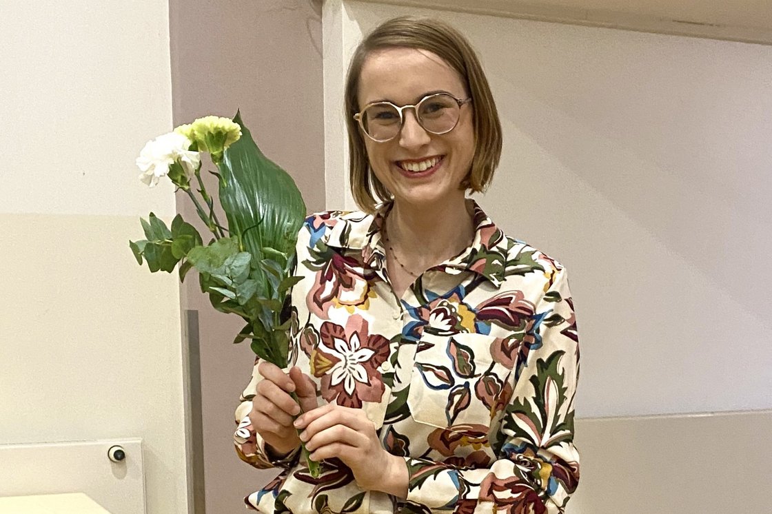 Eine lächelnde Frau in einer Blumenbluse und Brille hält einen kleinen Strauß weißer Blumen in einem Raum mit Holzboden.