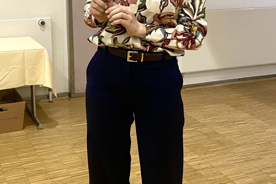 Eine lächelnde Frau in einer Blumenbluse und Brille hält einen kleinen Strauß weißer Blumen in einem Raum mit Holzboden.