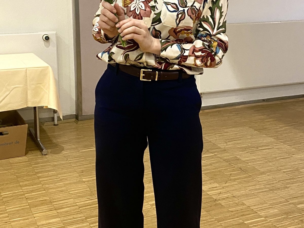 Eine lächelnde Frau in einer Blumenbluse und Brille hält einen kleinen Strauß weißer Blumen in einem Raum mit Holzboden.