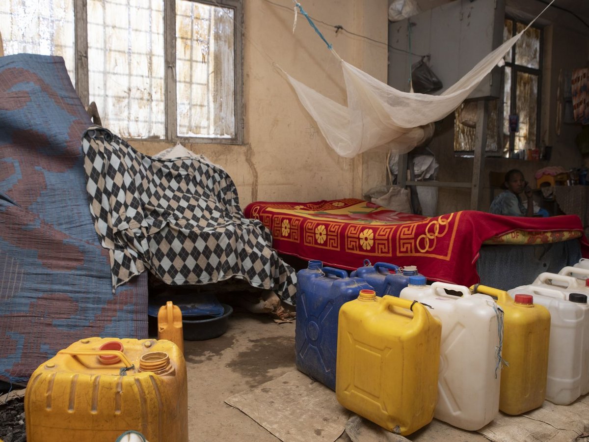 Einfaches Zimmer mit Bett, Moskitonetz und vielen Wasserkanistern am Boden. Ein Kind sitzt im Hintergrund.