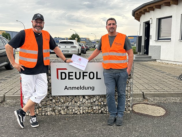 Zwei Männer in orangefarbenen Warnwesten stehen an einem 'DEUFOL Anmeldung' Schild, einer hält einen weißen Umschlag.