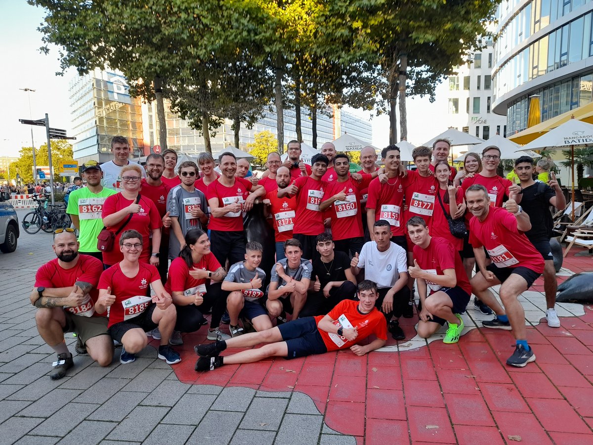 Große Gruppe von Don Bosco Sachsen Läufern in roten Shirts und Startnummern posiert mit Daumen hoch nach einem Lauf.