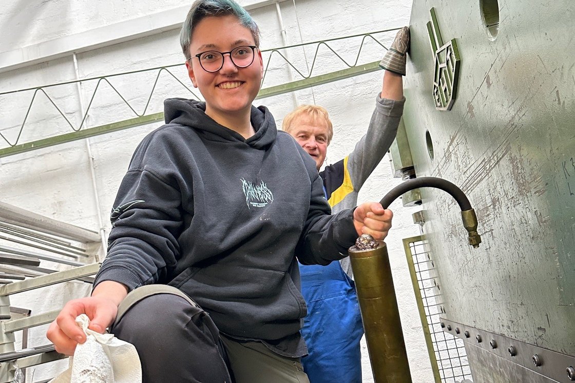 Eine junge Frau mit grauem Hoodie und Arbeitshose steht in einer Fabrikhalle und hält eine Gaskartusche in der Hand. Hinter steht ein Mann in Handwerkermontur. in einer Fabrikhalle 