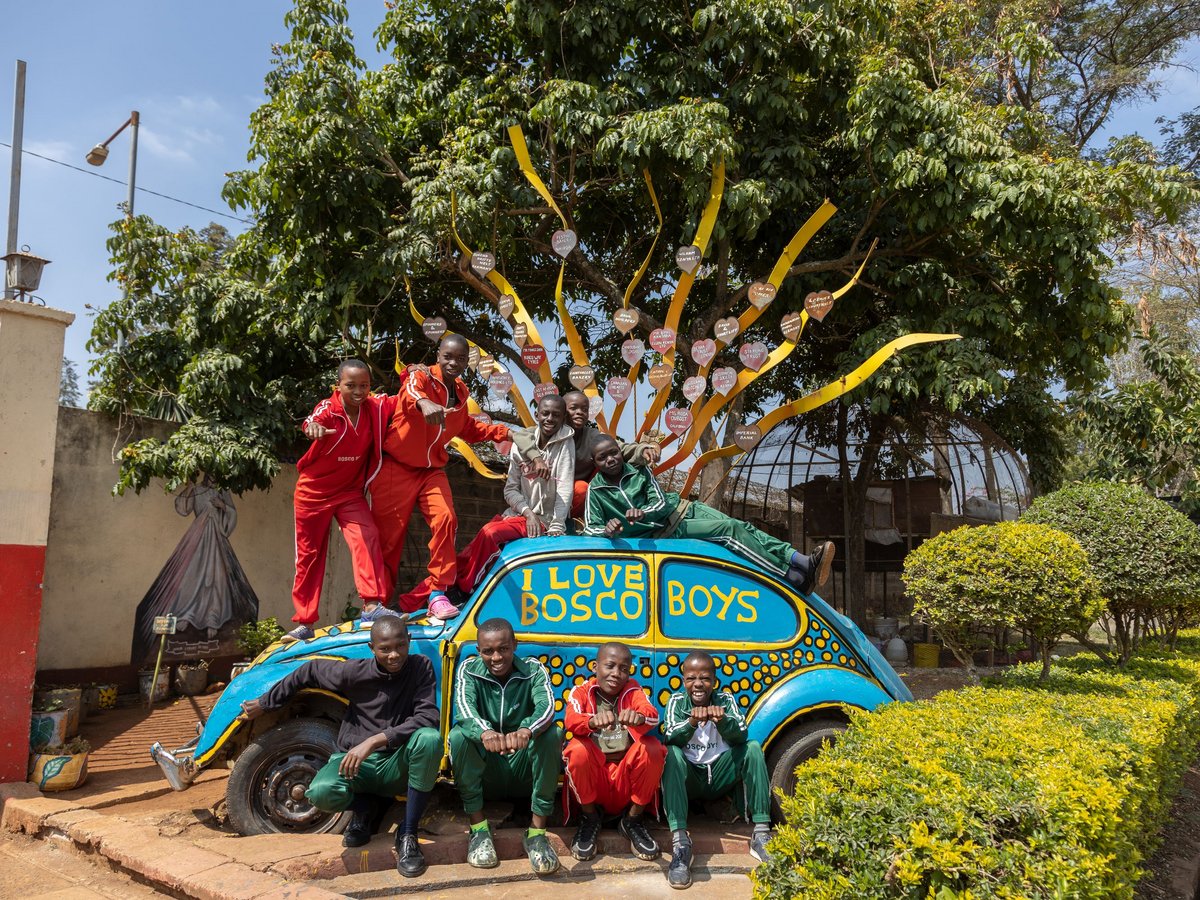 Lächelnde Jungen posieren auf und um ein blaues Auto mit „I LOVE BOSCO BOYS“, unter einem Baum mit Herzplaketten.