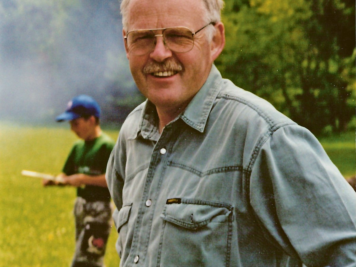 Lächelnder Mann mit Brille und Schnurrbart bei einem Outdoor-Treffen, mit einem Kind und Rauch im Hintergrund.