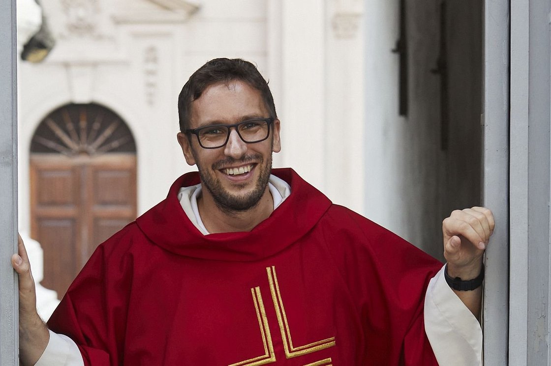 Ein lächelnder Priester in rotem Messgewand mit goldenem Kreuz steht in einem Türrahmen und schaut in die Kamera.