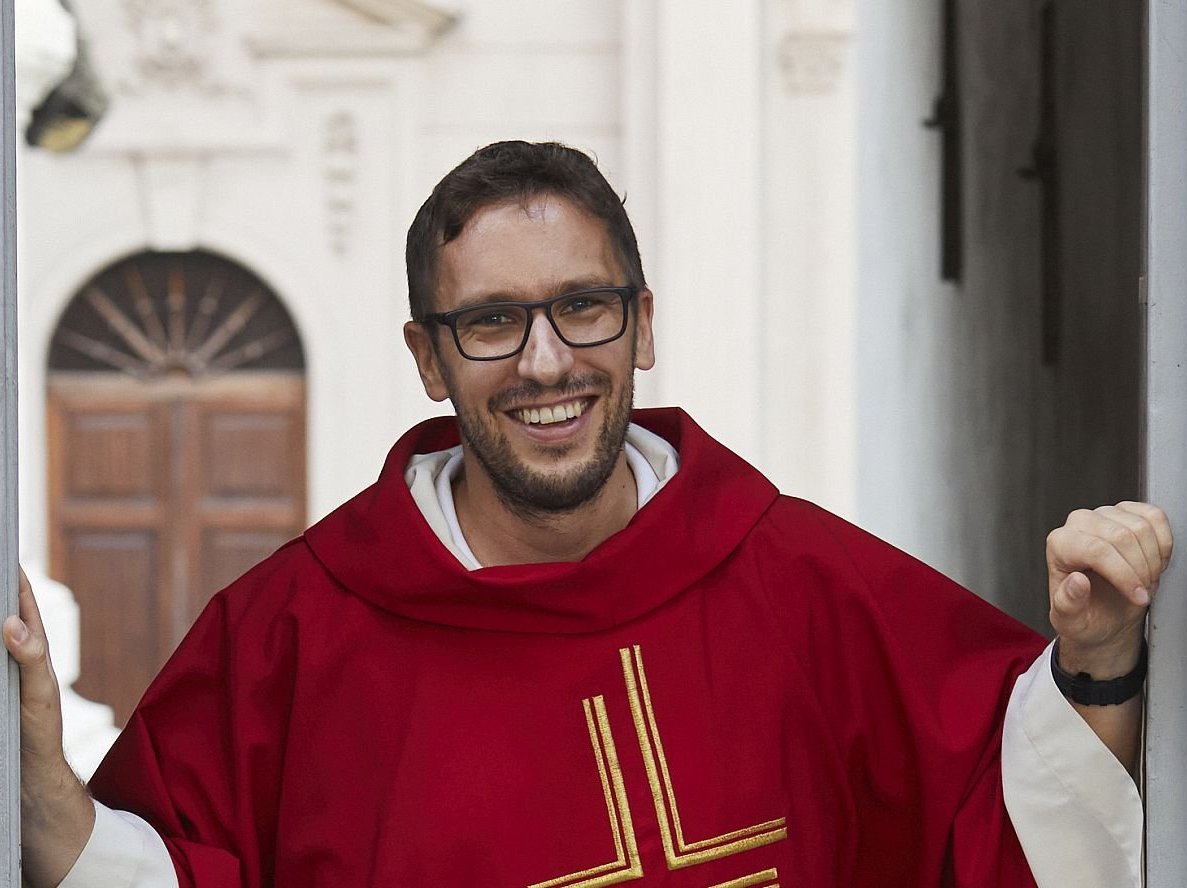 Ein lächelnder Priester in rotem Messgewand mit goldenem Kreuz steht in einem Türrahmen und schaut in die Kamera.