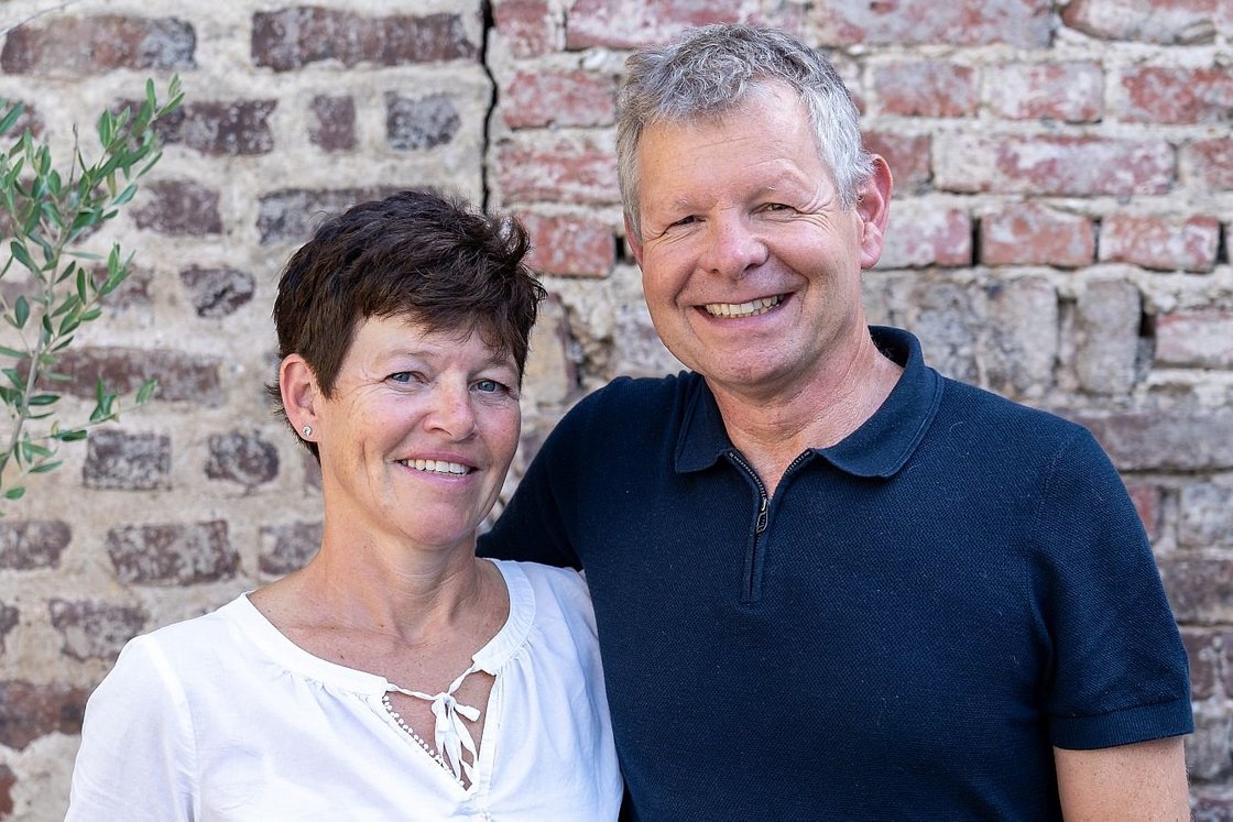 Lächelndes Paar, eine Frau mit kurzen braunen Haaren und ein Mann mit grauen Haaren, vor einer Ziegelmauer.