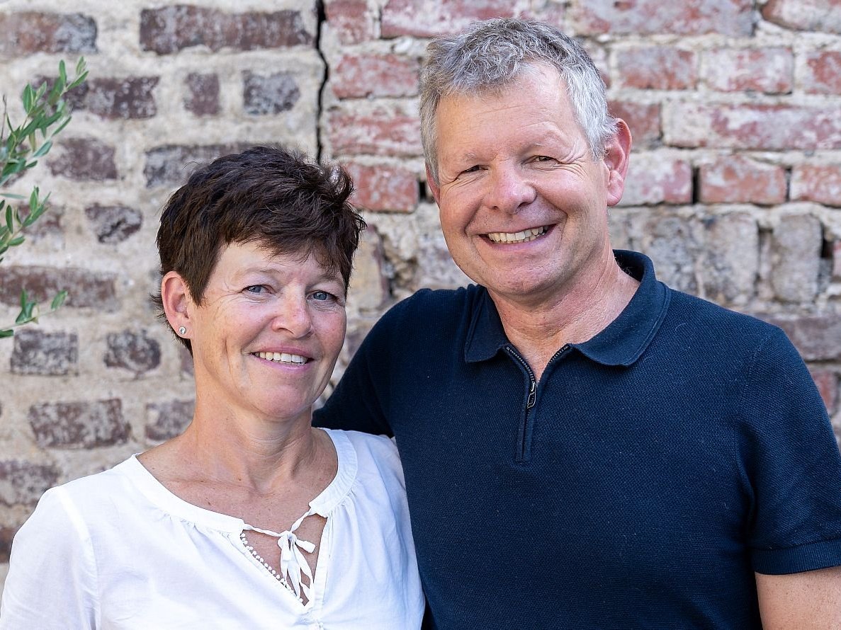 Lächelndes Paar, eine Frau mit kurzen braunen Haaren und ein Mann mit grauen Haaren, vor einer Ziegelmauer.