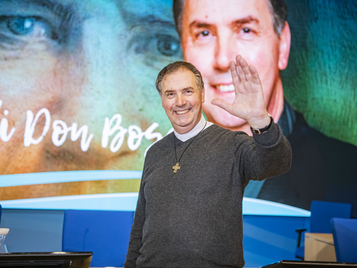 Lächelnder Priester in grauem Pullover und Kreuzkette winkt. Im Hintergrund Bildschirm mit „Don Bosco“ Schriftzug und Männergesicht.