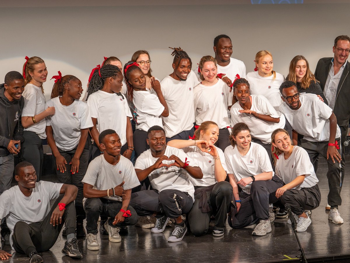 Eine fröhliche, diverse Gruppe junger Erwachsener in weißen T-Shirts und roten Bändern posiert lachend auf einer Bühne.