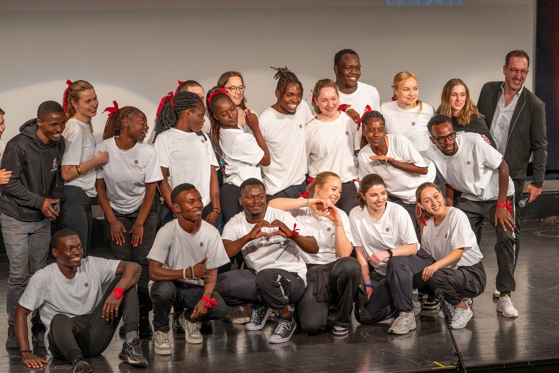 Eine fröhliche, diverse Gruppe junger Erwachsener in weißen T-Shirts und roten Bändern posiert lachend auf einer Bühne.