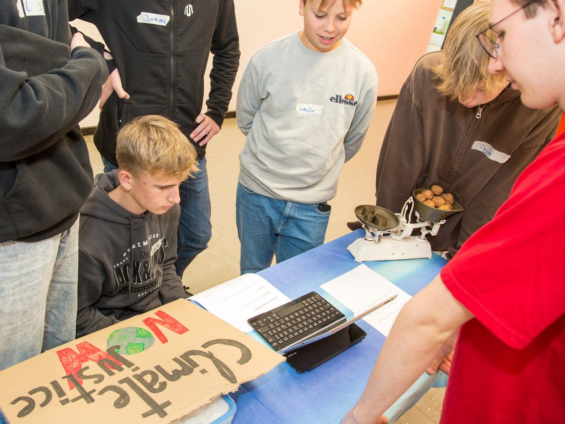 Schüler versammeln sich um einen Tisch mit einem 'Climate Justice NOW'-Schild, einer Waage und Walnüssen.