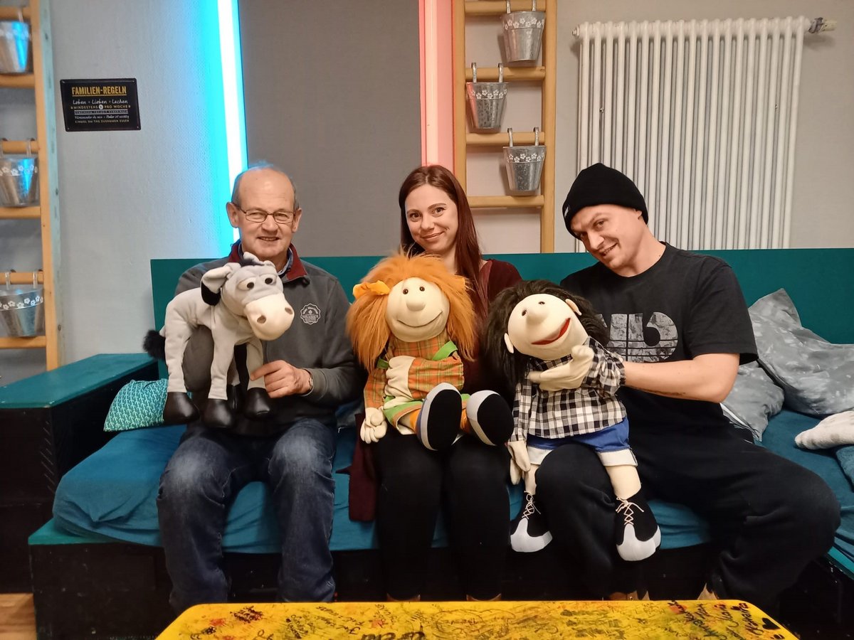 Drei Personen, zwei Männer und eine Frau, sitzen auf einem Sofa und lächeln mit je einer Handpuppe in die Kamera.
