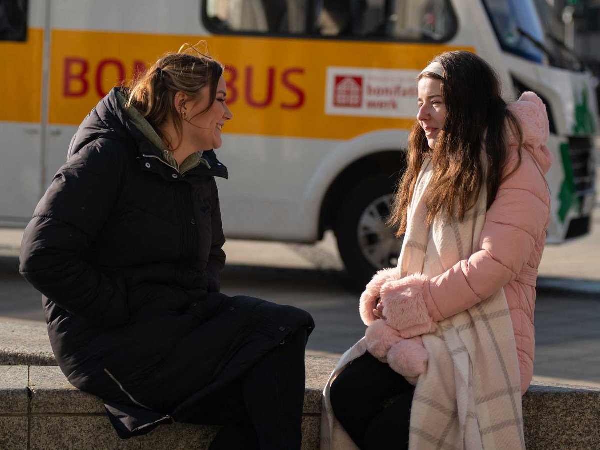 Zwei Frauen in Wintermänteln lächeln und unterhalten sich draußen auf einem Sims. Im Hintergrund ist ein gelber Bus mit "BOMBUS" zu sehen.