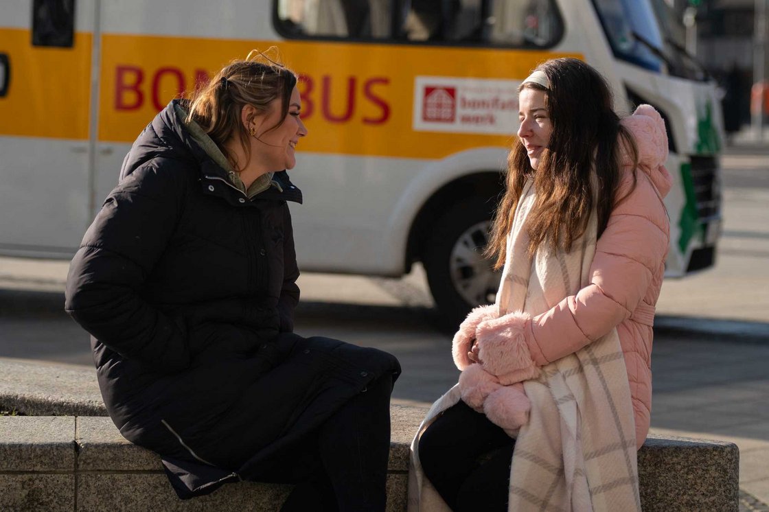 Zwei Frauen in Wintermänteln lächeln und unterhalten sich draußen auf einem Sims. Im Hintergrund ist ein gelber Bus mit "BOMBUS" zu sehen.