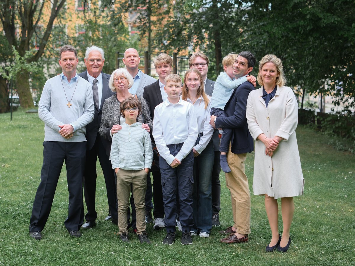 Großes Familienporträt mit mehreren Generationen, die lächelnd auf einer grünen Wiese vor Bäumen und Gebäuden posieren.