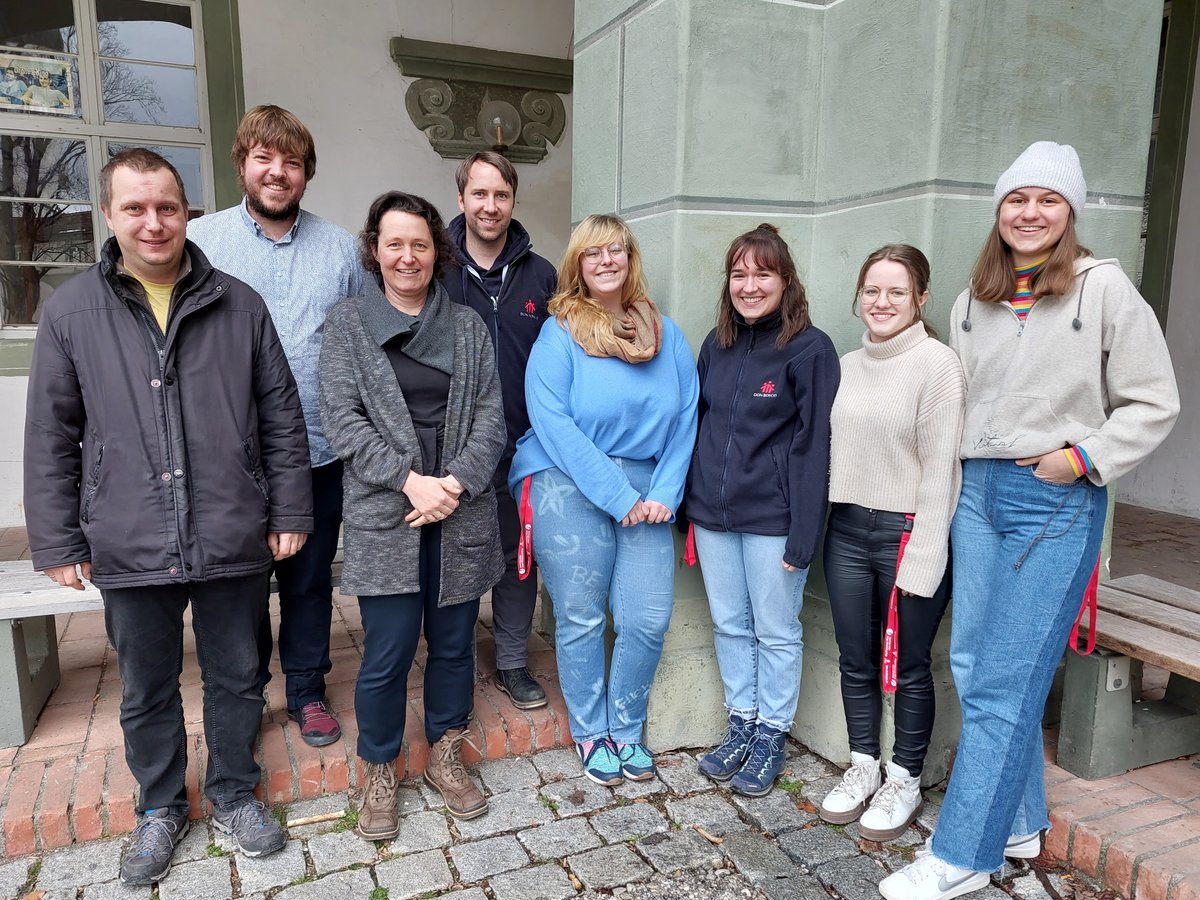 Neun lächelnde Personen (5 Frauen, 4 Männer) in Freizeitkleidung stehen draußen vor einem hellgrünen Gebäude.