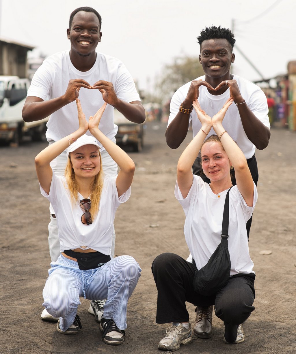 Vier lächelnde Menschen in weißen Shirts bilden Herz- und Hausformen mit ihren Händen auf einem Feldweg.