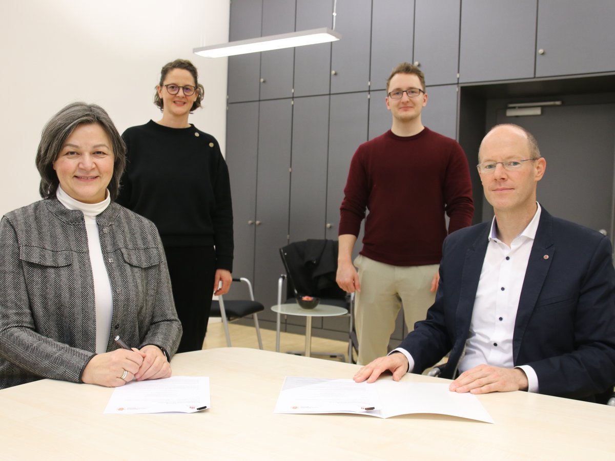 Vier Personen in einem Büro, zwei an einem Tisch mit Dokumenten sitzend, zwei stehend dahinter, alle lächeln.
