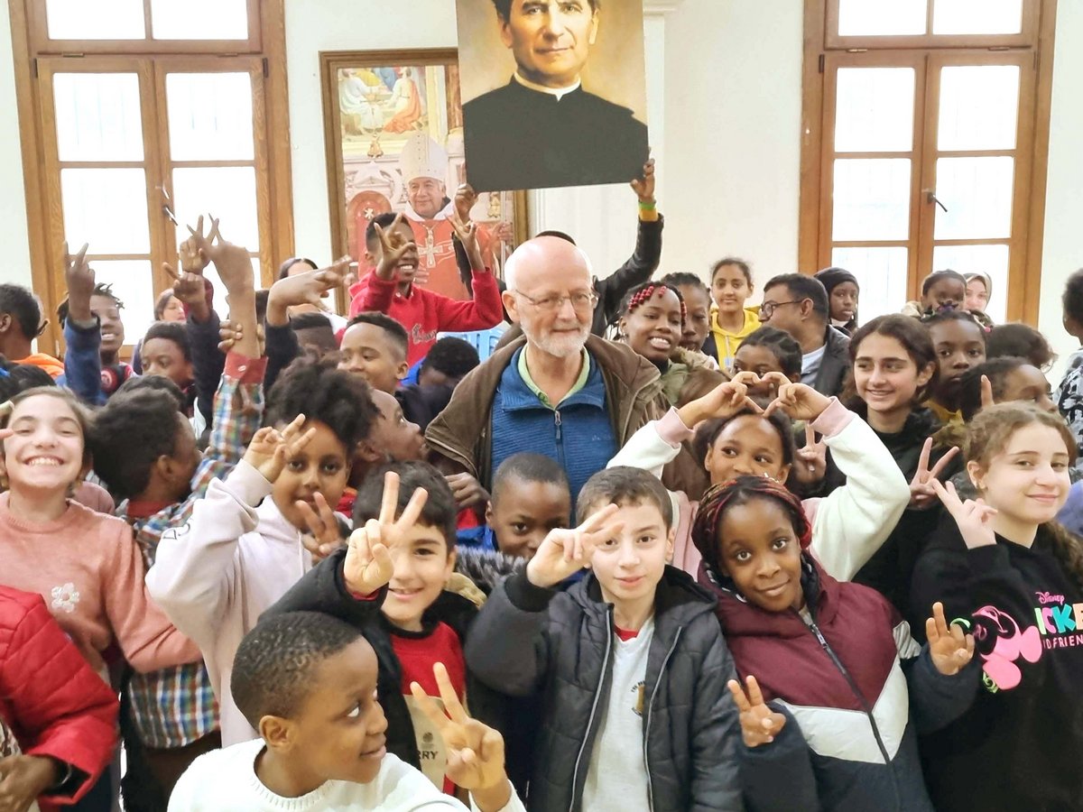Eine vielfältige Gruppe fröhlicher Kinder und Erwachsener posiert drinnen, viele machen Friedenszeichen, mit einem Don-Bosco-Porträt im Hintergrund.