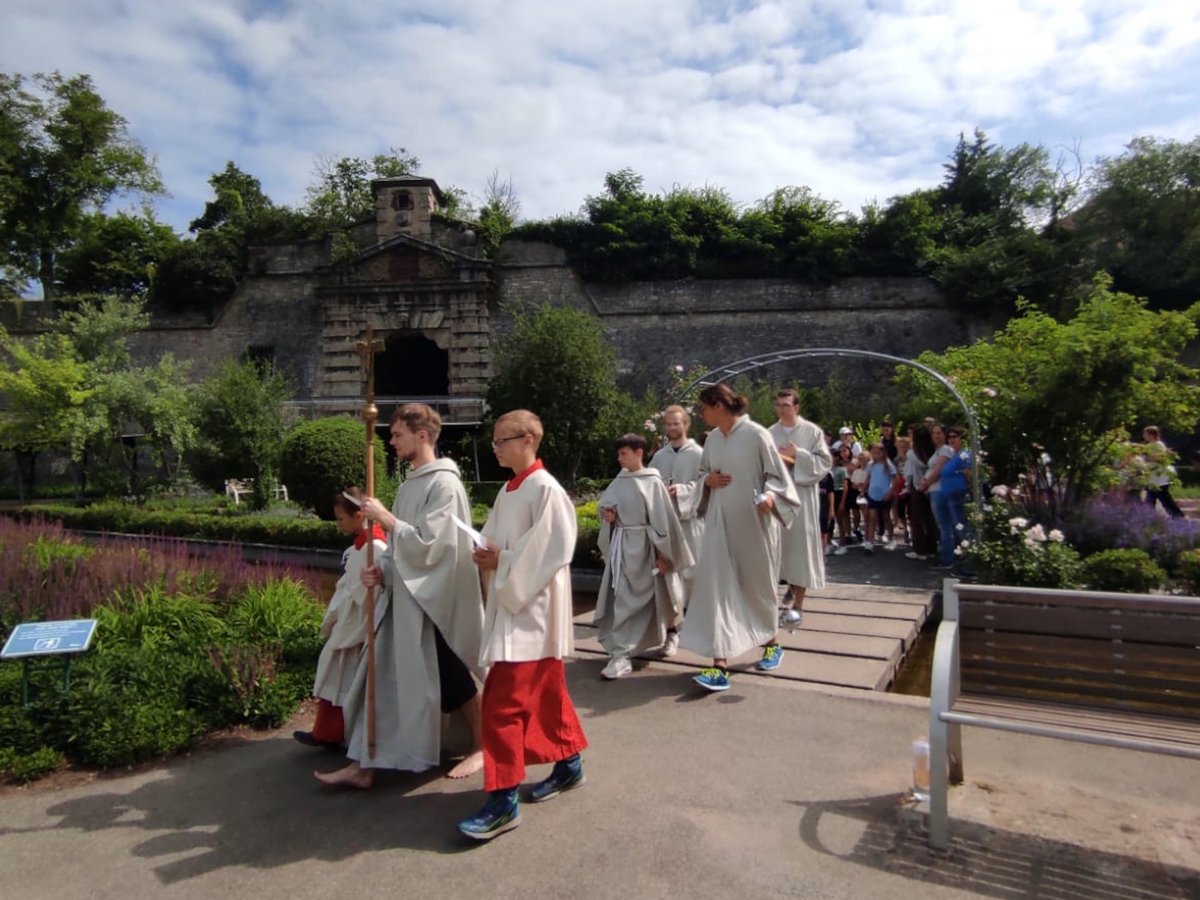 Prozession in Gewändern, teils barfuß, teils mit Turnschuhen, vor alter Steinmauer und Garten.