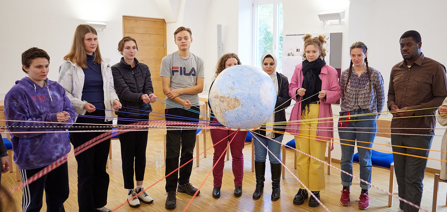 Vielfältige junge Menschen im Kreis halten farbige Fäden, die sie mit einem Globus verbinden, Symbol für globale Gemeinschaft.
