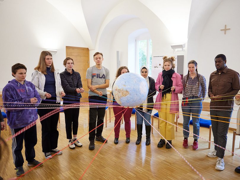Vielfältige junge Menschen im Kreis halten farbige Fäden, die sie mit einem Globus verbinden, Symbol für globale Gemeinschaft.