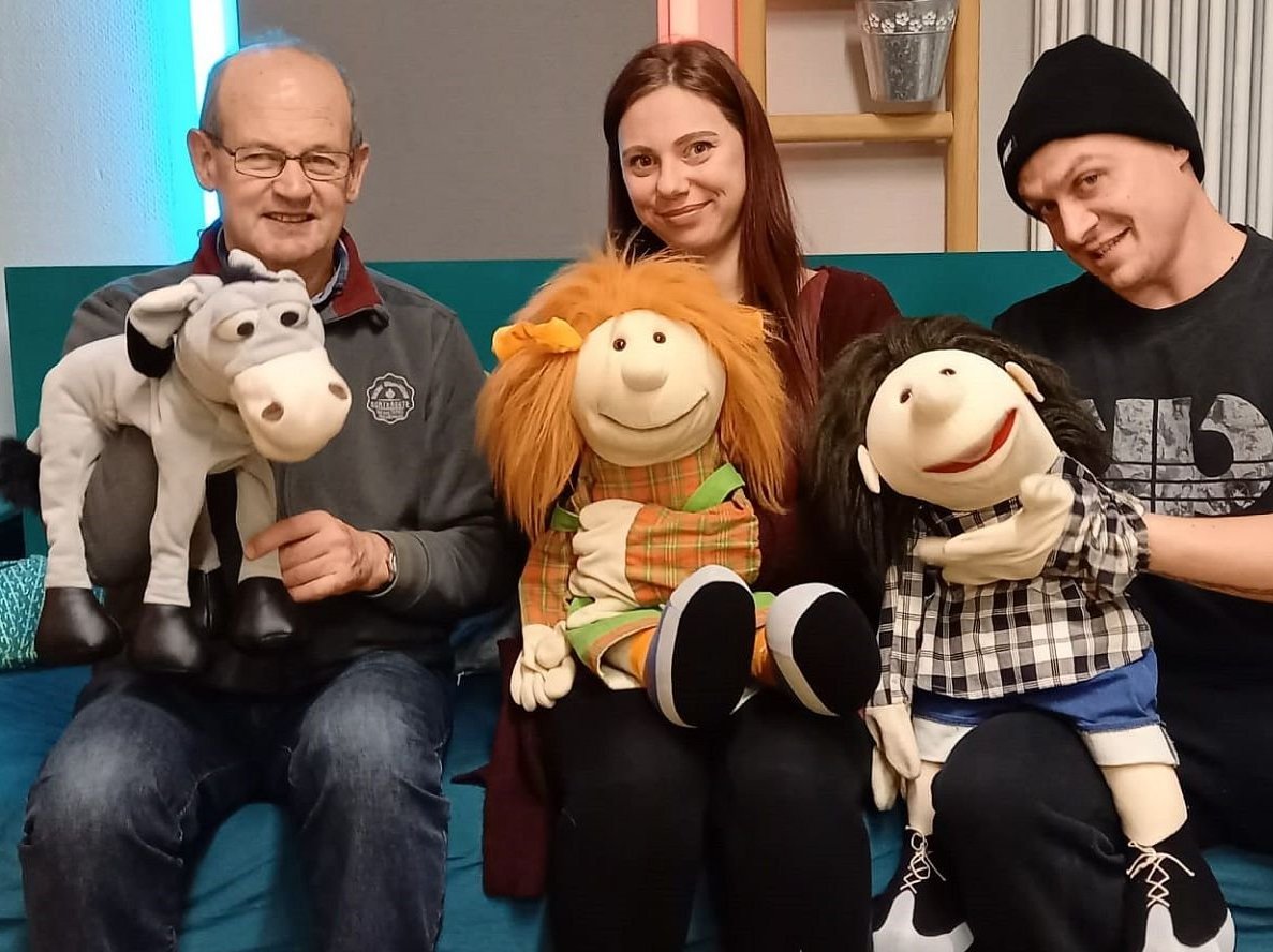 Drei Personen und ihre lächelnden Handpuppen: Esel, Mädchen mit roten Haaren, Junge mit dunklen Haaren auf einer Couch.