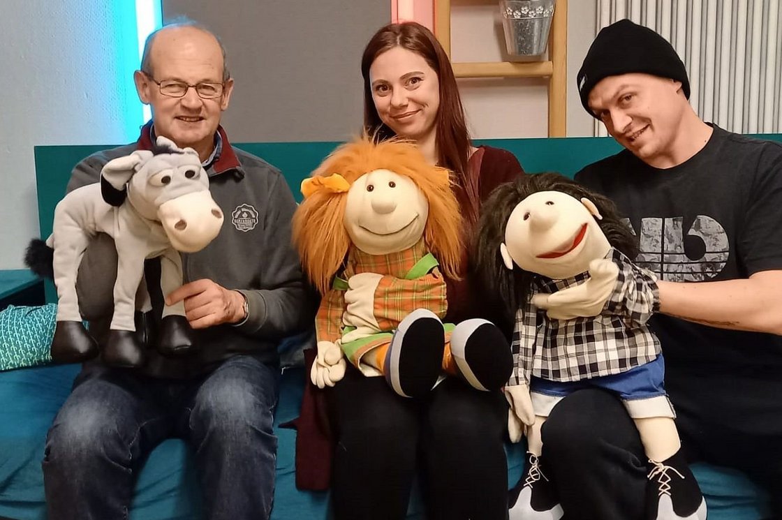 Drei Personen und ihre lächelnden Handpuppen: Esel, Mädchen mit roten Haaren, Junge mit dunklen Haaren auf einer Couch.