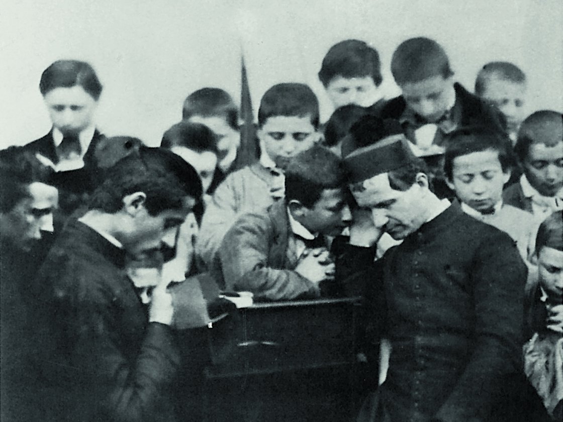 Monochromes Foto: Ein Priester in Soutane spricht mit einem knienden Jungen mit gefalteten Händen, umgeben von Jungen, beim Religionsunterricht.