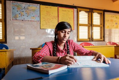 Eine junge Schülerin im rot gestreiften Hemd sitzt am blauen Schreibtisch, liest ein Buch und markiert etwas.