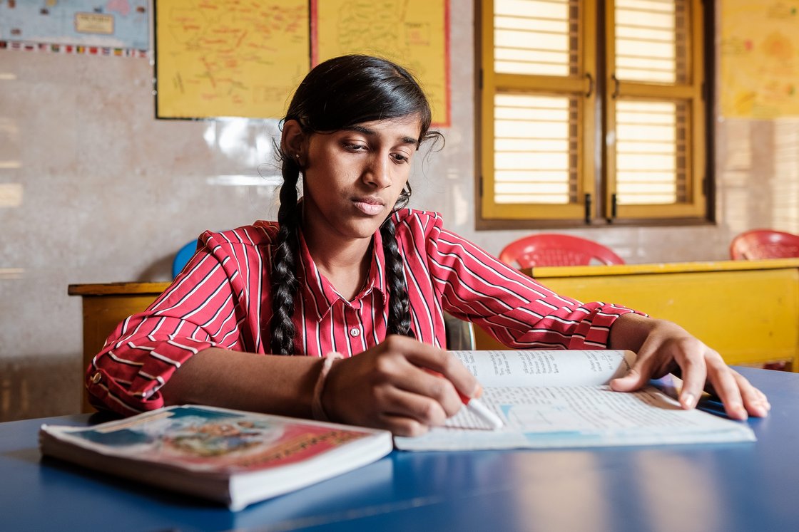 Eine junge Schülerin im rot gestreiften Hemd sitzt am blauen Schreibtisch, liest ein Buch und markiert etwas.