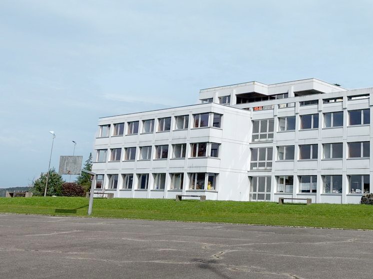 Weißes, modernes Gebäude mit vielen Fenstern hinter einem Asphaltplatz und Basketballkorb, umgeben von Bäumen und Gras.