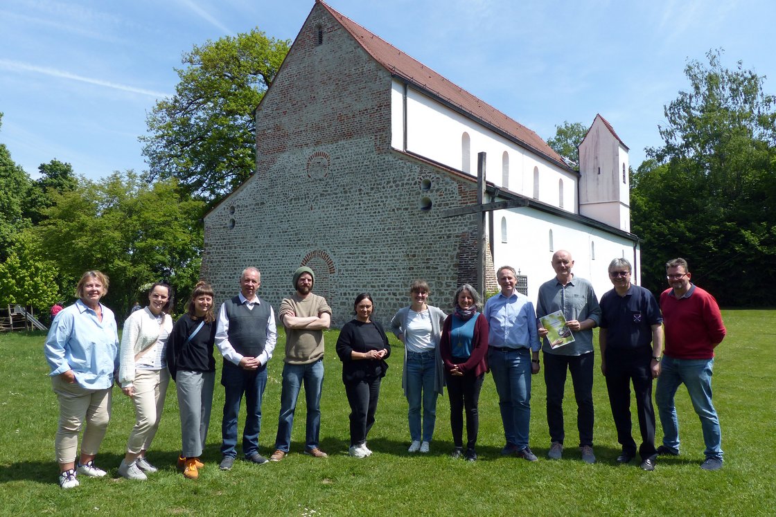 Gruppe von 12 Personen steht auf einer Wiese vor einer alten Kirche mit Holzkreuz unter blauem Himmel.
