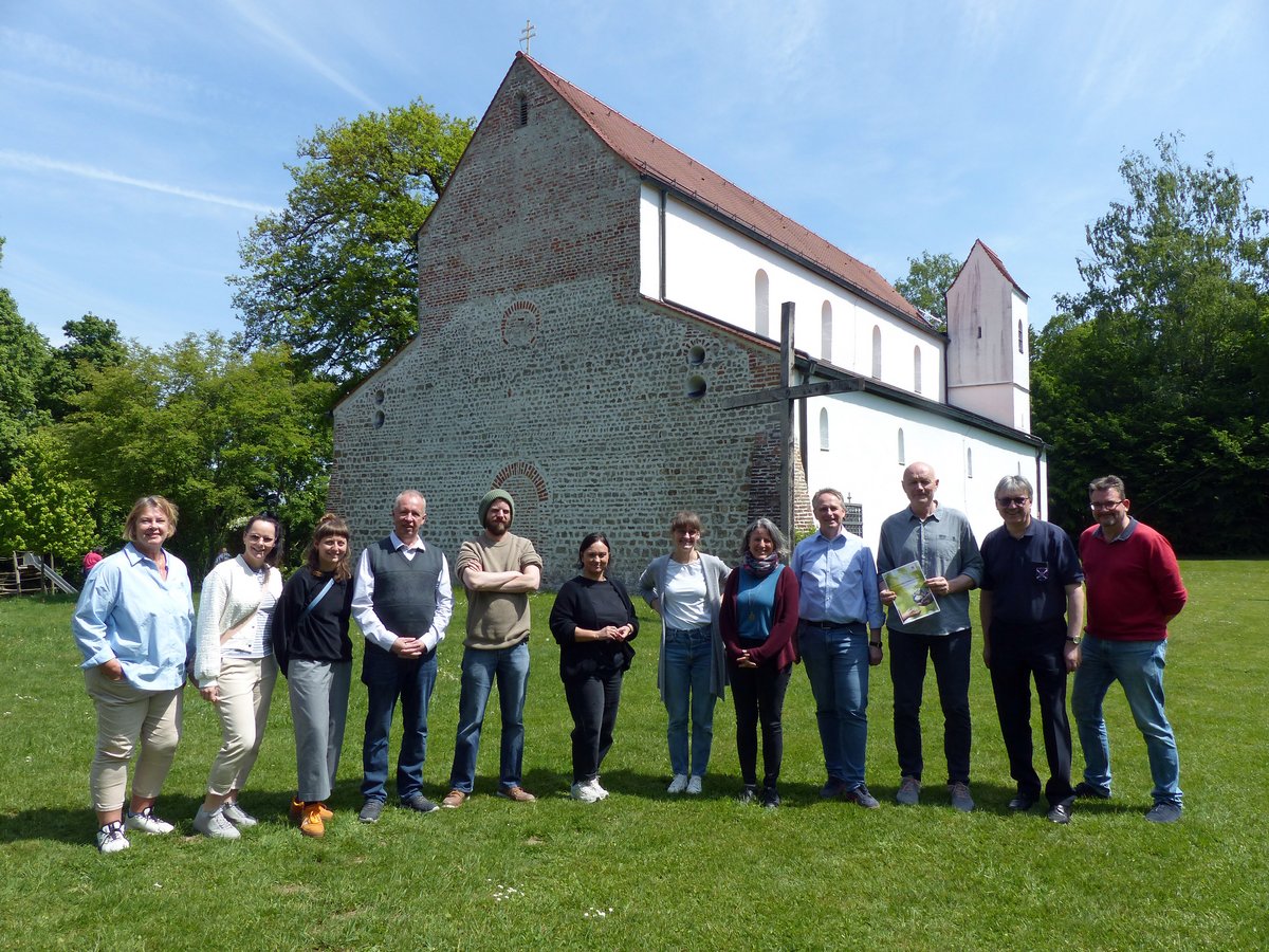 Gruppe von 12 Personen steht auf einer Wiese vor einer alten Kirche mit Holzkreuz unter blauem Himmel.